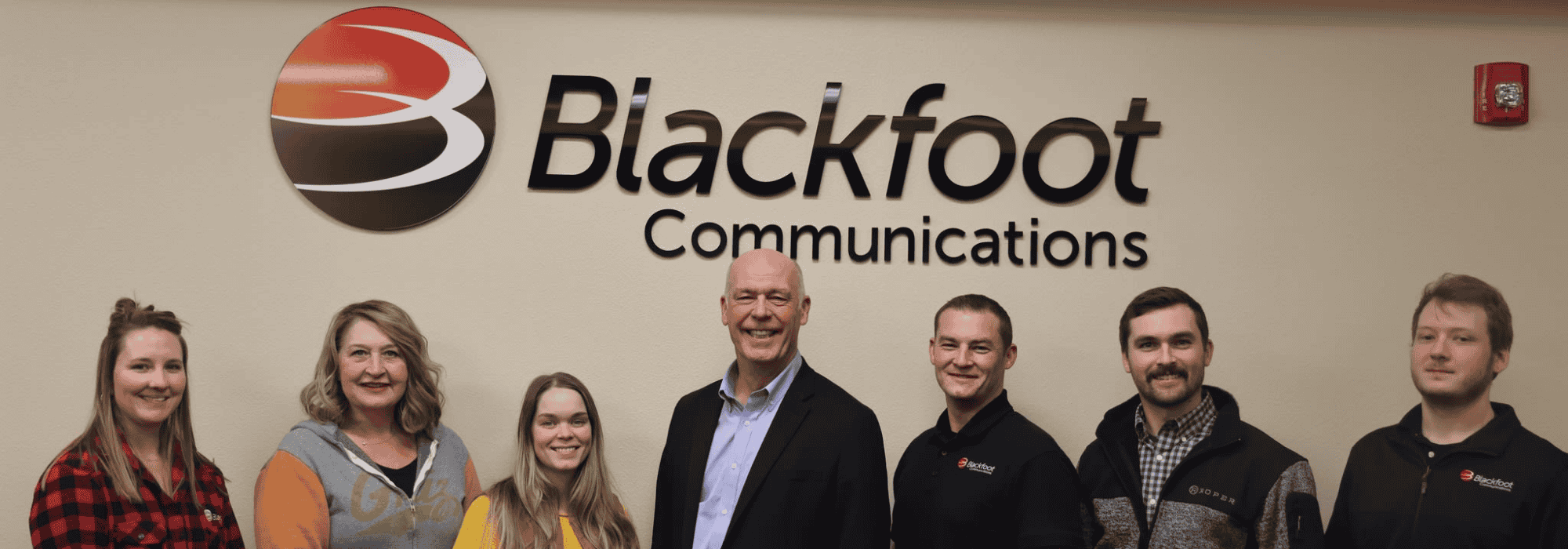 Six Blackfoot Communications team members and our governor smiling in front of a branded wall sign at the office.